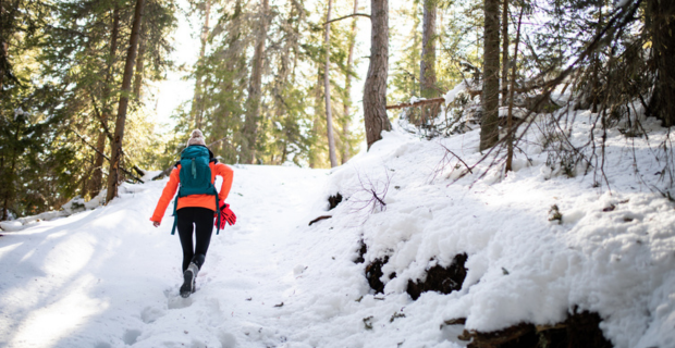 Une femme, vêtue d’un manteau orange avec des gants attachés à son sac à dos bleu, monte la colline à grands pas. La neige recouvre le sol. Le soleil est visible à travers les arbres de la forêt.