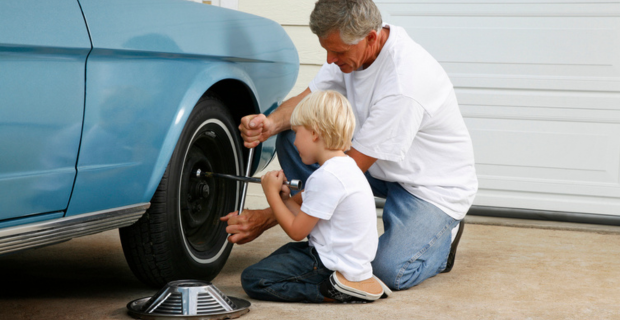 Un homme portant un chandail blanc et des jeans montre à un jeune enfant portant un chandail blanc et des jeans comment utiliser un cric pour les pneus. Un enjoliveur de roue est au sol à côté de l’enfant. Devant eux se trouve une magnifique voiture classique bleu clair.