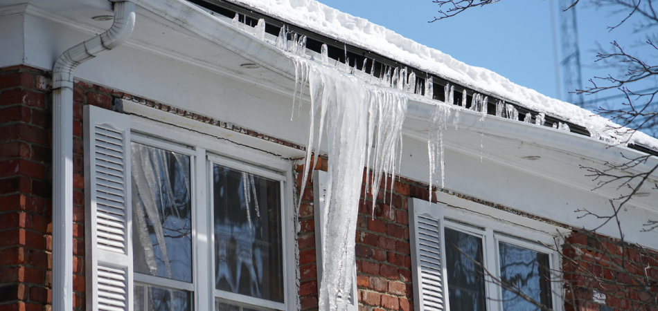 Glaçons suspendus au bord d’un toit enneigé d’une maison en briques avec des volets blancs, sous un ciel bleu éclatant d’hiver.  