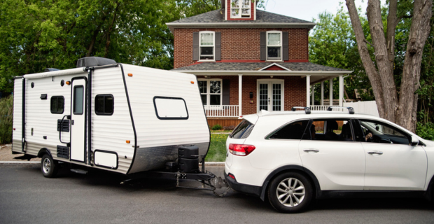 Une voiture blanche à quatre portes quitte la maison en tirant une caravane blanche pour partir à l’aventure! À l’arrière-plan, on aperçoit une maison en briques rouges avec une véranda panoramique et un grand arbre dans la cour avant.