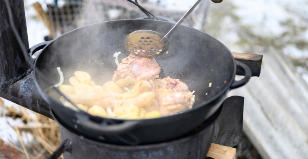De la viande et des légumes sont cuits dans une casserole de fonte ovale au-dessus d’un feu en plein air. On voit la vapeur s’élever des aliments savoureux, qui sont remués par une personne à l’aide de sa main gantée.