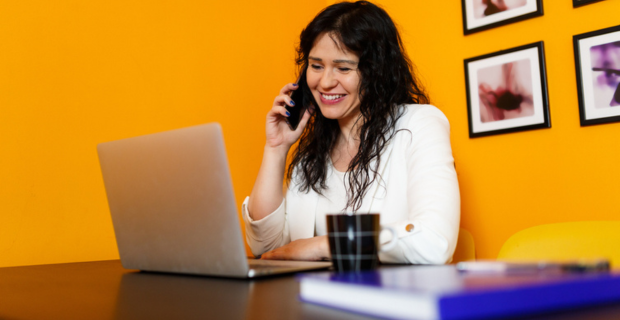 Une jeune femme parle au téléphone devant son portable dans son bureau ayant des murs orange et quelques tableaux accrochés derrière elle. 