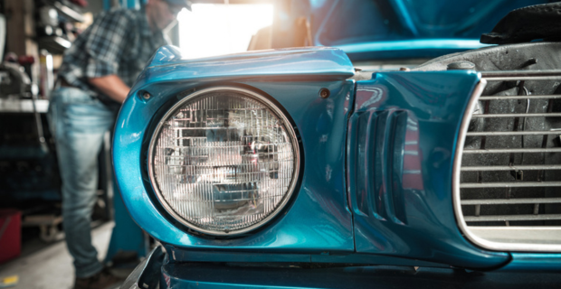 Gros plan d’une voiture de collection d’un bleu époustouflant avec point de mire sur le  phare avant double. Un homme vêtu d’une chemise à carreaux, d’un chapeau et d’un jean travaille sur la magnifique voiture en arrière-plan