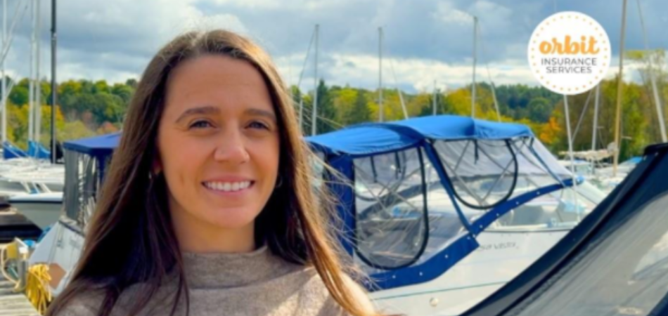 Une personne aux cheveux longs se tient debout sur un quai et parle d’assurance marine. Elle sourit et porte un chandail beige tandis que plusieurs bateaux sont amarrés derrière elle.
