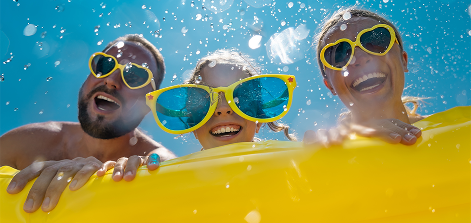 Scène d'été amusante avec trois personnes derrière un flotteur de piscine jaune, portant des lunettes de soleil et des lunettes de protection colorées, avec des éclaboussures d'eau et un ciel bleu vif.