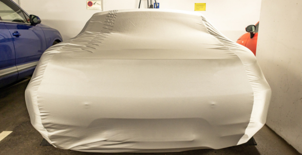Une voiture de collection dans un garage entre une voiture bleue et une voiture orange. Elle est couverte d’une housse de protection de qualité de couleur havane.