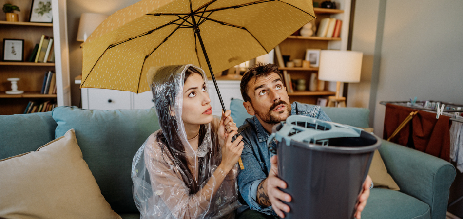 Un homme assis à côté de sa compagne sur le canapé de leur maison, tenant un seau pour récupérer l’eau qui s’échappe du toit, tandis que sa compagne tient un parapluie.