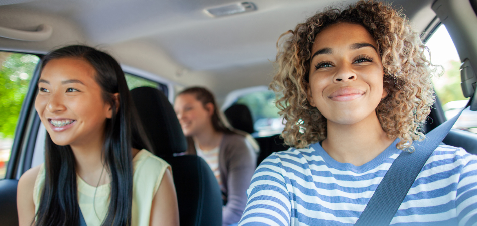 Une jeune au volant d’une voiture avec deux passagères, toutes attachées, voyageant ensemble par une journée ensoleillée.