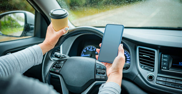 Photo mettant en gros plan une personne qui conduit avec une main qui tient un café sur le volant et l’autre qui tient un cellulaire