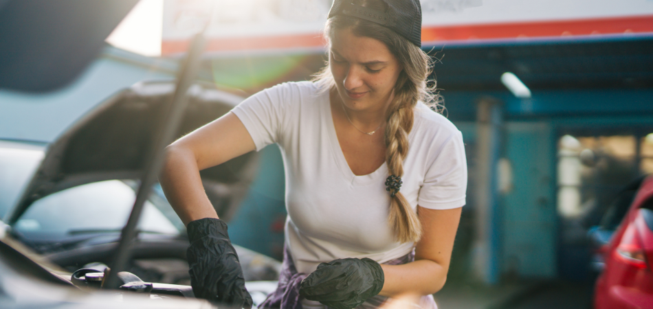 Une jeune femme regardant sous le capot d’un véhicule afin d’effectuer l’entretien printanier de sa voiture classique et de la préparer à prendre la route.
