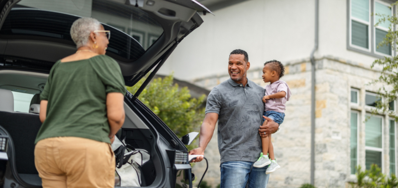 Une personne aux cheveux gris courts décharge son véhicule électrique tandis qu’une autre personne tient un enfant et recharge le véhicule. La voiture est stationnée dans un quartier résidentiel et elles discutent de l’assurance fondée sur l’utilisation.   