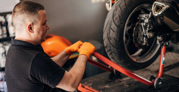 Un homme portant une chemise noire avec un col et des gants de travail orange clair hisse un cric orange foncé avec la moto sur l’élévateur. Seul le pneu est visible avec du métal brillant. La moto est levée à la hauteur des yeux de l’homme qui travaille sur la machine. 