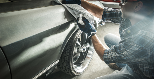 Un homme porte un jean, une chemise à carreaux, un chapeau et des lunettes de sécurité. Il a des gants bleus et utilise une serviette blanche pour faire briller sa précieuse voiture de collection. Devant la voiture argentée sur laquelle il travaille se trouve une superbe voiture de collection rouge.