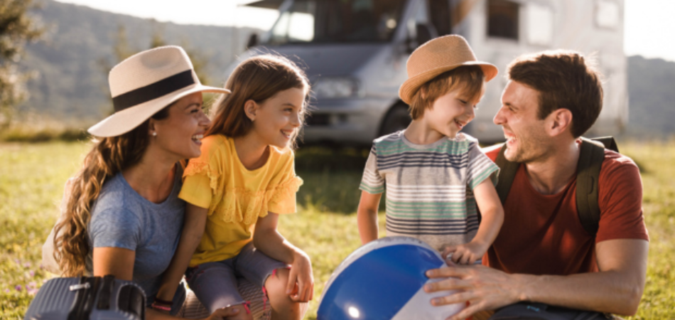Une famille de quatre personnes en voyage en VR est assise sur l’herbe devant un VR, vêtue de vêtements d’été décontractés; un enfant tient un ballon de plage.