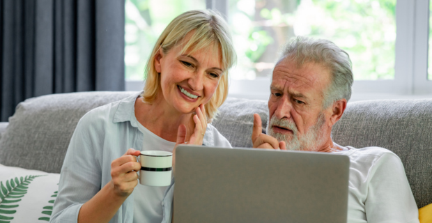 Image d’un couple d’âge moyen assis sur un sofa avec un ordinateur portable sur les genoux. La femme, qui tient une tasse à café blanche avec une rayure bleue, est assise avec la main qui touche sa mâchoire. L’homme fait un geste vers le haut avec son index et semble engagé dans une conversation ou poser une question. 