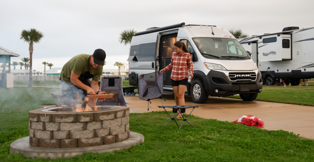 Un homme et des femmes debout près d’un foyer. Il y a un camping-car en arrière-plan.