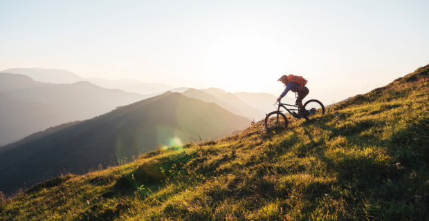 Un cycliste portant un sac à dos et descendant la côte d’une montagne verdoyante. Au loin, une belle chaîne de montagnes s’efface progressivement.