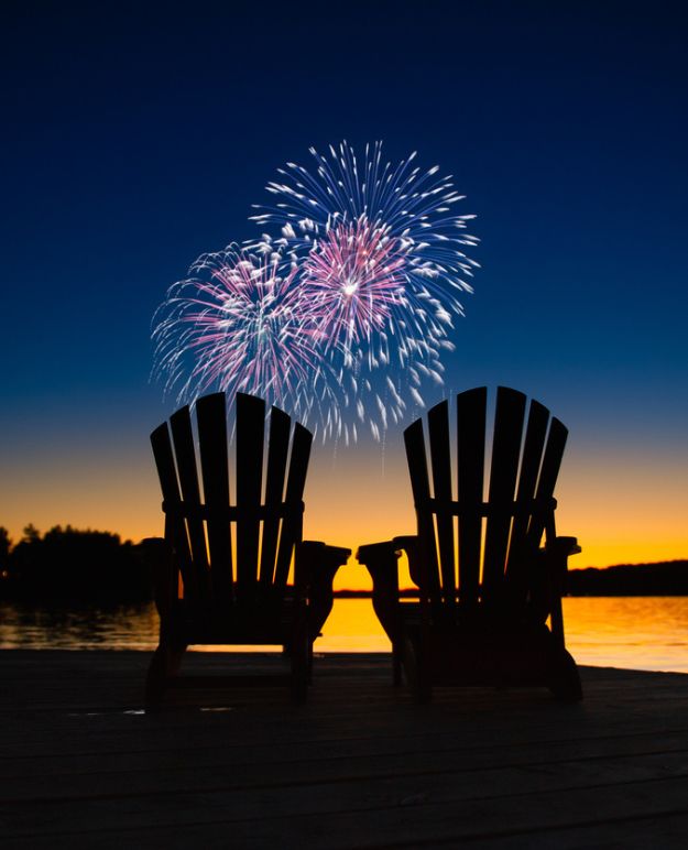 Feux d'artifice sur un lac immobile alors que la soirée tourne au crépuscule. Treescape silhouette le lac avec deux chaises Adirondack au premier plan.