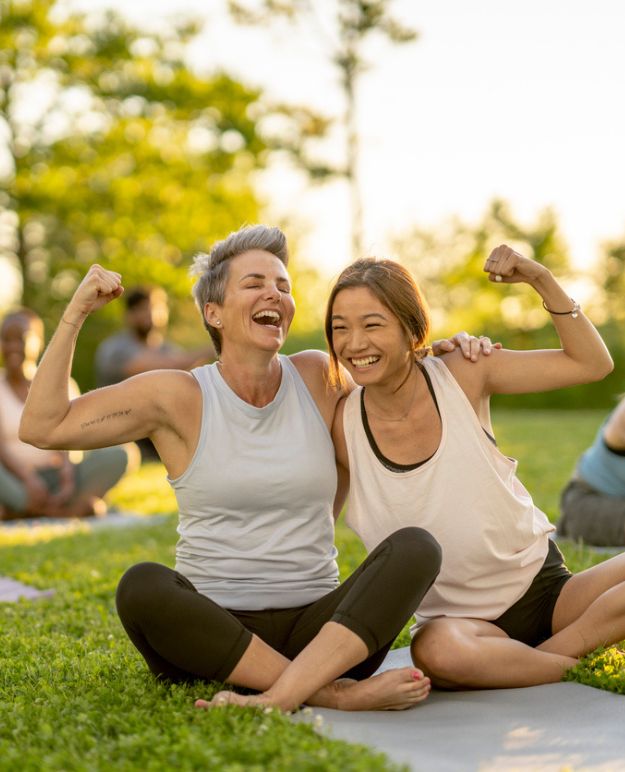 Deux femmes assises en tailleur dans un parc faisant du yoga un soir d'été en Alberta. Ils rient, les bras autour des épaules l'un de l'autre, tandis qu'ils exhibent leurs biceps en fléchissant.
