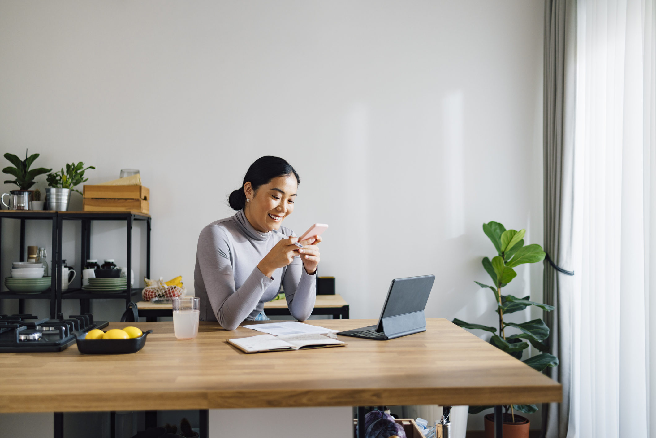 Femme assise à une table de cuisine avec un ordinateur portable et un document, souriant à son téléphone