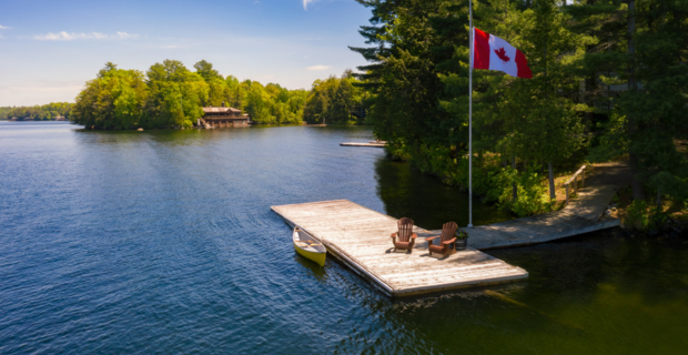 Image d’un lac aux eaux parfaitement calmes par une belle journée ensoleillée. On peut voir un quai et un canot. Un drapeau canadien est fièrement arboré. Le chalet d’un voisin est visible de l’autre côté du lac.