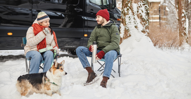 Un couple assis sur des chaises dans un camping d’hiver avec leur chien devant leur VR.  