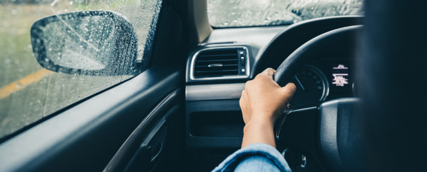 Une photo de quelqu’un qui conduit une auto sous la pluie, prise à partir du siège arrière. On ne voit que sa main droite qui tient le volant. 