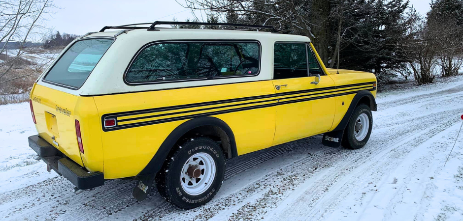 SUV International Harvester Scout II jaune vif des années 1970 garé sur une allée en gravier enneigée, vu depuis l’angle arrière gauche.