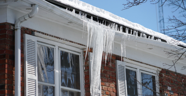 Glaçons suspendus au bord d’un toit enneigé d’une maison en briques avec des volets blancs, sous un ciel bleu éclatant d’hiver.  
