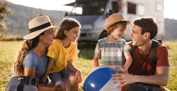 Une famille de quatre personnes en voyage en VR est assise sur l’herbe devant un VR, vêtue de vêtements d’été décontractés; un enfant tient un ballon de plage.