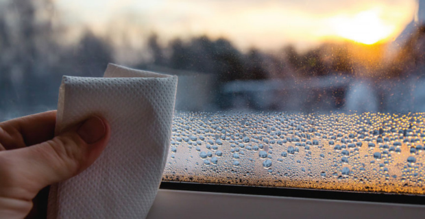 Gros plan sur la main d’une personne utilisant un essuie-tout pour sécher les gouttes de condensation sur une fenêtre en verre par un froid matin d’hiver au lever du soleil.
