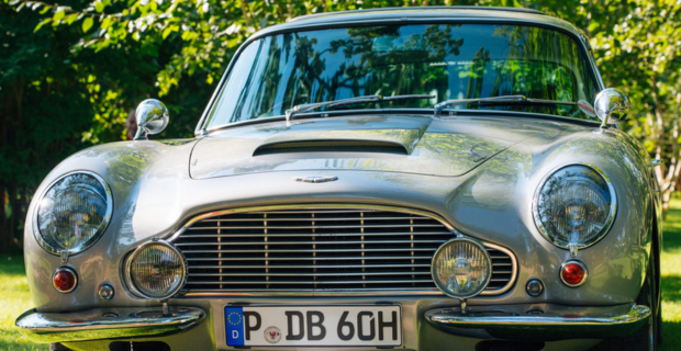 Gros plan de la calandre et de l’avant de la fabuleuse DB5. La peinture de couleur gris métallique est splendide et reflète le soleil. En arrière-plan, on peut voir le feuillage des arbres.