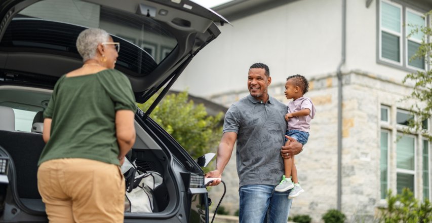 Une personne aux cheveux gris courts décharge son véhicule électrique tandis qu’une autre personne tient un enfant et recharge le véhicule. La voiture est stationnée dans un quartier résidentiel et elles discutent de l’assurance fondée sur l’utilisation. 