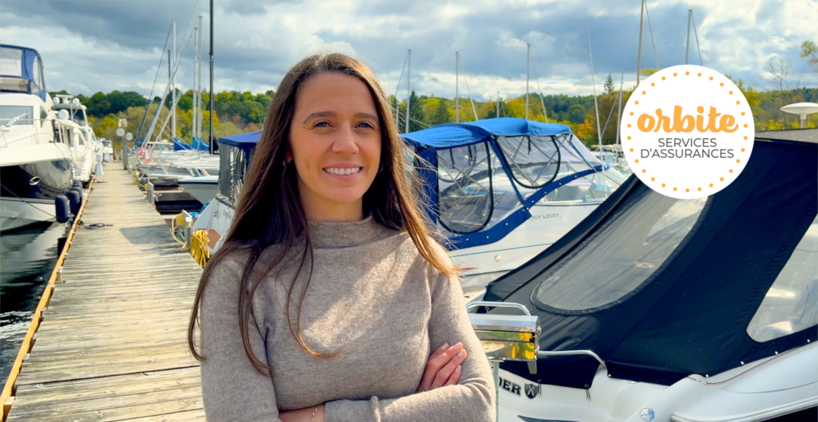 Une personne aux cheveux longs se tient debout sur un quai et parle d’assurance marine. Elle sourit et porte un chandail beige tandis que plusieurs bateaux sont amarrés derrière elle.